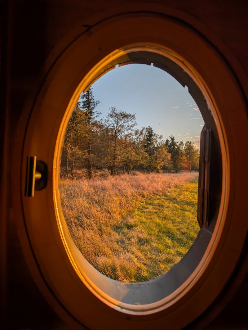 Pohled kulatým oknem tiny house U Lesa na louku ve večerním světle